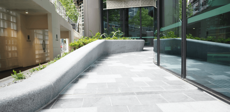 A sleek curved natural stone bench and light grey granite pavers along a glass-fronted building in a landscaped courtyard